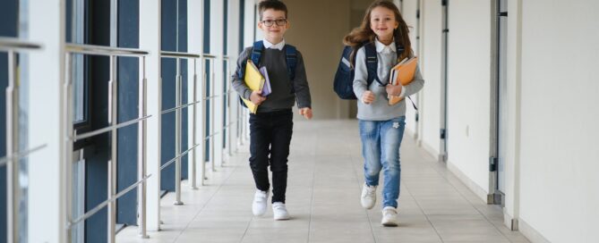 Two smiling schoolchildren with backpacks and books walking down a school corridor