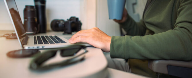 Person working on a laptop in a camper van, holding a mug, with camera equipment and sunglasses nearby.