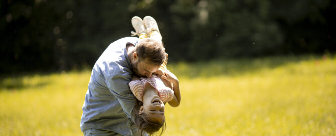 Father playing with his daughter outdoors in a sunny park, holding her upside down while they both laugh and enjoy a fun, carefree moment together.