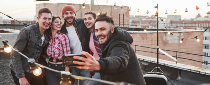Group of young friends smiling and taking a selfie on a rooftop, surrounded by string lights and a cityscape in the background.