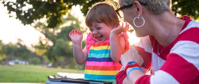 A mother and her young daughter sitting outdoors, smiling and interacting joyfully, with a tablet computer placed on the table in front of them.