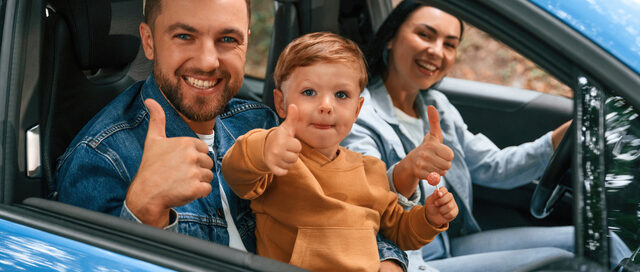 Happy family with young child giving thumbs up while sitting in a car together