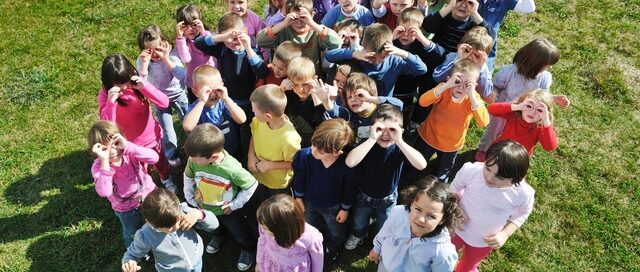 Large group of children and adults gathered together in a circle on grass, viewed from above