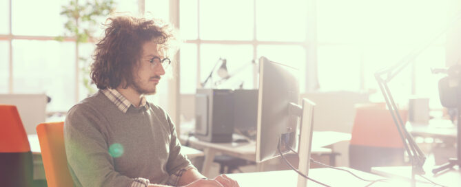 A man with curly hair and glasses sits at a desk working on a computer in a bright office with large windows.