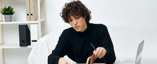 Person with curly brown hair in black sweater writing in notebook at wooden desk with laptop and white shelving unit with plants in background.