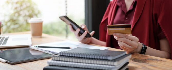 Person in red shirt holding smartphone and credit card at desk with laptop, notebooks, and coffee cup, making an online purchase.