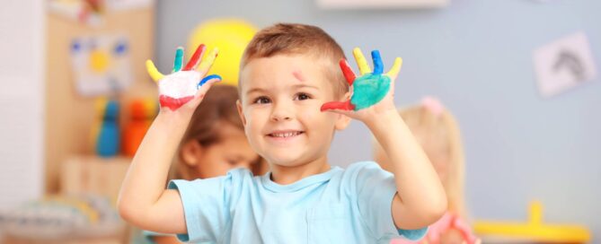 a child in a classroom with multiple colours of paint on his hands, smiling