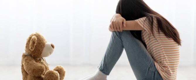 a sad child sitting down with a teddy bear with her head buried into crossed arms and hunched legs