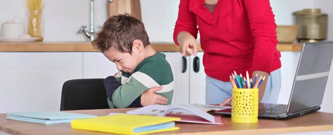 an angry child with folded arms turned away from a woman who is pointing at the books on the table