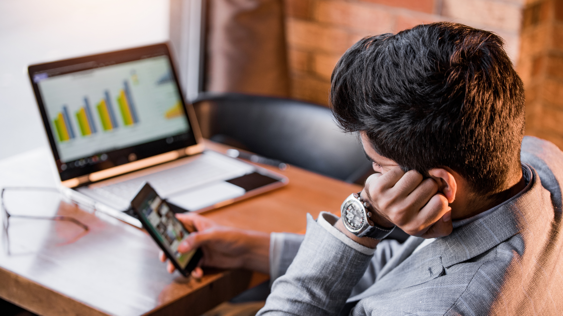 A man procrastinating on his phone sitting at his desk with an open laptop